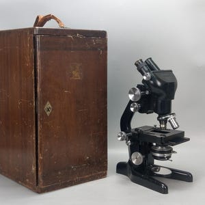 Op de afbeelding: Een zwarte metalen binoculaire microscoop met verstelbare focusknoppen, een mechanische tafel en een ronde spiegelvoet, naast een hoge, rechthoekige, verweerde bruine houten opbergkist met een lederen handvat aan de bovenzijde.