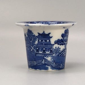 May include: A small, white ceramic planter with a blue and white willow pattern. The planter features a pagoda, trees, and a fence.