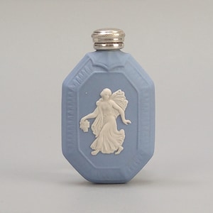 Op de afbeelding: Lichtblauwe, achthoekige parfumfles met een zilverkleurige dop. De fles heeft een wit bas-reliëf van een klassieke figuur met bloemen. Het ontwerp doet denken aan antieke Wedgwood-stukken.