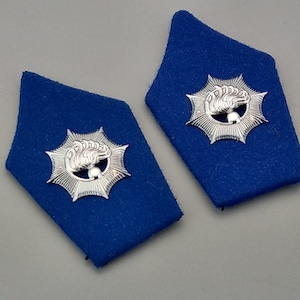May include: Two blue shoulder boards with a silver emblem featuring a stylized design. The emblem has a central element surrounded by a starburst pattern. The boards are a deep blue color.