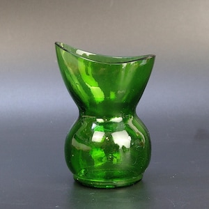 Könnte beinhalten: Eine kleine grüne Glasvase mit einer ungewöhnlichen Sanduhrform. Die Vase hat einen abgerundeten Boden und eine ausgestellte Oberseite, wodurch eine unverwechselbare Silhouette entsteht. Die leuchtende grüne Farbe und das glänzende Finish tragen zur visuellen Attraktivität bei. Geeignet für kleine Blumen.
