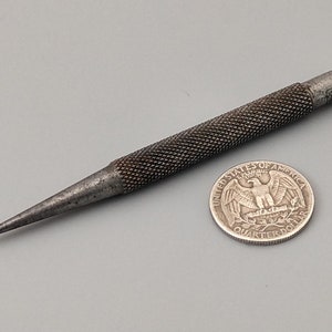 May include: A silver metal punch tool with a textured handle and a pointed tip. The tool is about the size of a US quarter.