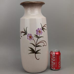 May include: A tall, off-white ceramic vase with a brown rim and floral design. The vase features hand-painted purple flowers and green leaves on a slender, cylindrical body. The vase is approximately 25 cm tall.