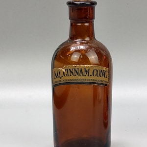 May include: A vintage brown glass bottle with a cork stopper. The bottle has a label with the text "AQ. CINNAM. CONC." The bottle is likely an antique apothecary or medicine bottle.