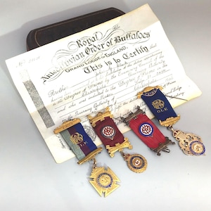 May include: An assortment of antique fraternal order medals and a certificate from the Royal Antediluvian Order of Buffaloes. The medals feature gold-toned metal, colorful ribbons, and intricate designs. The certificate is aged with visible text.