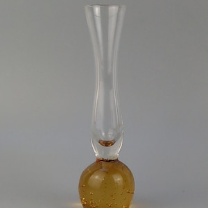 Könnte beinhalten: Eine klare Glas-Blumen-Vase mit einem bauchigen, bernsteinfarbenen Sockel. Der Sockel hat kleine Luftblasen, und die Vase verjüngt sich zu einem schlanken Hals. Die Vase ist etwa 20 cm hoch, geeignet für eine einzelne Blume oder ein kleines Bouquet.