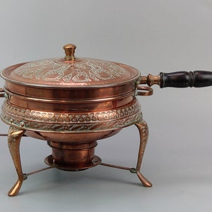 May include: A copper chafing dish with an ornate design and a wooden handle. The dish sits on a stand with three legs.
