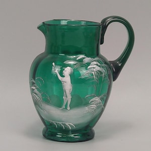 May include: Green glass jug with a handle and spout. The jug has a white etched design of a person and palm trees. The jug is a decorative item.