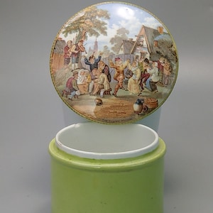 Puede incluir: Un tarro redondo de cerámica verde lima con interior y borde blancos. La tapa presenta una ilustración detallada de una escena de pueblo con gente bailando y tocando música. El texto "THE VILLAGE WEDDING" es visible en la tapa.