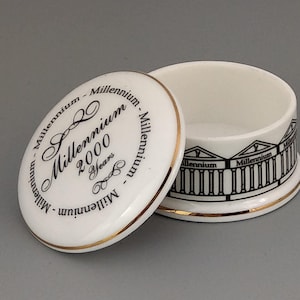 Puede incluir: Caja de cerámica blanca con un borde dorado. La tapa tiene una inscripción negra que dice "Millennium 2000 2 Years Millennium - Millennium - Millennium - Millennium". La base de la caja tiene un diseño en blanco y negro de tres edificios con la palabra "Millennium" inscrita en cada uno.