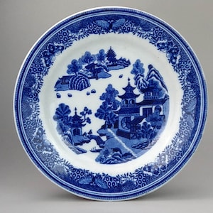 Peut inclure: Une assiette en céramique blanche avec un motif bleu et blanc représentant un paysage chinois avec des pagodes et des arbres. L'assiette a une bordure bleue et blanche avec un motif floral.