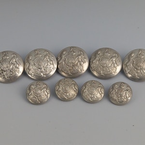 Może przedstawiać: Zestaw dziewięciu srebrnych metalowych guzików o różnych rozmiarach. Każdy guzik ma szczegółowy wytłoczony wzór herbu z koroną, lwami i innymi elementami heraldycznymi. Ułożone na jasnoszarym tle.