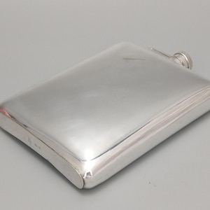 Op de afbeelding: Een rechthoekige, zilverkleurige flacon met een afgeronde bovenkant en een kleine, ronde dop. De flacon heeft een glad, reflecterend oppervlak en een eenvoudig, elegant ontwerp. De flacon is waarschijnlijk gemaakt van metaal en is ontworpen om vloeistoffen in te bewaren.