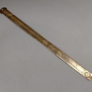 Könnte beinhalten: Ein antikes Messlineal aus Messing mit geätzten Markierungen und einem kleinen Loch an einem Ende. Das Lineal hat Zentimeter- und Bruchteilmessungen sowie ein dekoratives Emblem. Das Lineal hat eine warme Goldfarbe.
