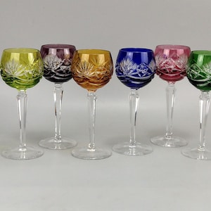 Könnte beinhalten: Sechs farbige Kristall-Weingläser. Jedes Glas hat eine andere Farbe: Gelb, Lila, Orange, Blau, Rosa und Grün. Die Kelche haben ein geschliffenes Design und die Stiele sind klar.