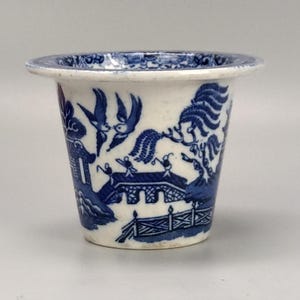 May include: A small, white ceramic planter with a blue and white willow pattern. The planter is shaped like a cone and has a wide rim.