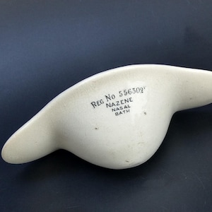 May include: Off-white ceramic nasal bath with a curved, ergonomic design. The text "REG No 556302" and "NAZENE NASAL BATH" are printed on the surface. The object is on a dark background.