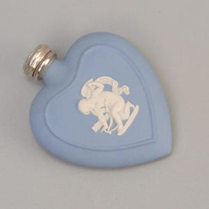 Op de afbeelding: Een hartvormige, lichtblauwe parfumfles met een zilveren dop. De fles heeft een wit bas-reliëf ontwerp van klassieke figuren. Een decoratief item om tentoon te stellen of te gebruiken.