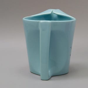 May include: A light blue ceramic mug with a unique angular design. The mug features a square handle and a folded rim. The mug's body is a solid, pale blue color, and the handle is a matching shade. The mug is a simple yet stylish piece.