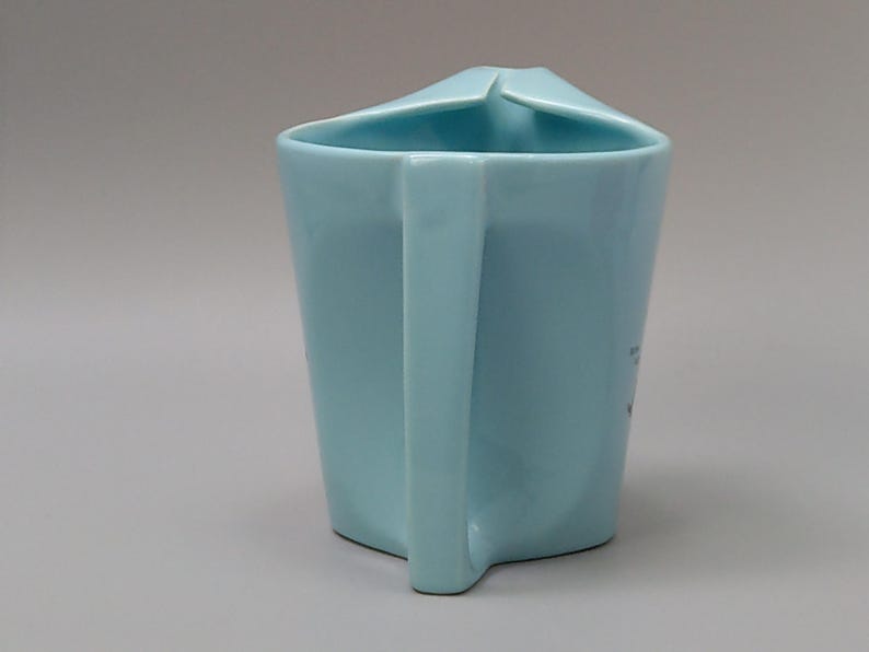 May include: A light blue ceramic mug with a unique angular design. The mug features a square handle and a folded rim. The mug's body is a solid, pale blue color, and the handle is a matching shade. The mug is a simple yet stylish piece.