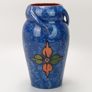 May include: A blue ceramic vase with a stylised floral design in orange, green and black. The vase has two handles that curve out from the sides.