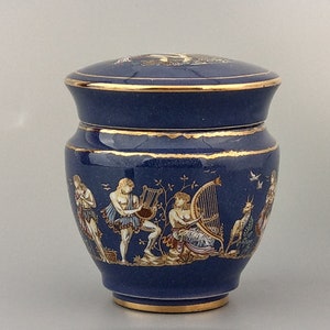 Può includere: Un vaso in ceramica blu e oro con un coperchio, che presenta una scena di persone che suonano musica e una capra. Il vaso ha un bordo dorato e un design dettagliato.