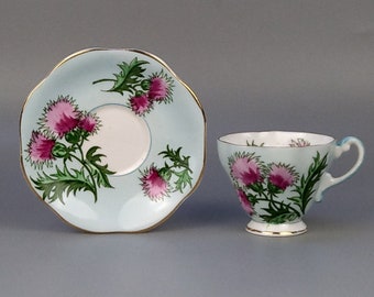 May include: A light blue teacup and saucer set with a floral design featuring pink thistles and green leaves.