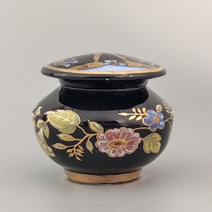 Può includere: Un piccolo vaso in ceramica nera con coperchio, decorato con motivi floreali dorati e colorati. Il vaso ha un corpo rotondo e un bordo leggermente svasato.
