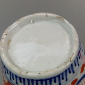 May include: The base of a white porcelain cup with blue and red painted designs. The cup has a white glaze and is decorated with a floral pattern.