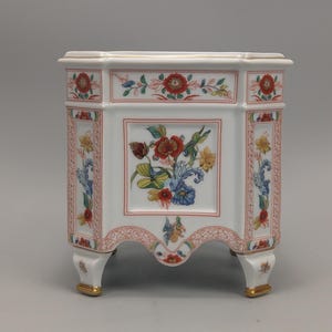 Pode incluir: Uma caixa decorativa de cerâmica branca com um design floral. A caixa tem uma forma octogonal com uma tampa e quatro pernas. O design floral inclui flores vermelhas, amarelas e azuis com folhas verdes. As bordas são douradas.