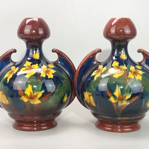 Könnte beinhalten: Zwei Keramikvasen mit blauer und grüner Glasur und gelben Blumendesigns. Die Vasen haben einen runden Körper und zwei Griffe an den Seiten.