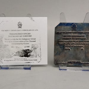 May include: A white paper certificate of authenticity for a Royal Crown Derby Porcelain Co. Ltd. Madagascan Tortoise, alongside a corresponding metal printing plate with reversed text, both displayed on clear plastic stands against a neutral background.