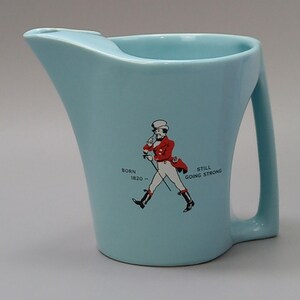 May include: A light blue ceramic pitcher with a handle and a pouring spout. The pitcher features a graphic of a man in a red coat and the text "Born 1820 - Still Going Strong."