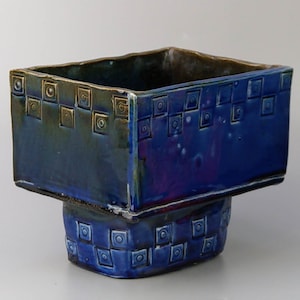 May include: A blue ceramic planter with a square top and a square base. The planter has a geometric pattern of squares and circles around the top and bottom.