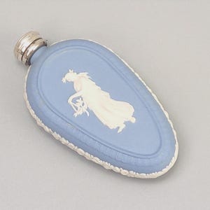 Op de afbeelding: Een lichtblauwe fles met een wit reliëf van een klassieke figuur. De fles heeft een zilverkleurige dop en een witte decoratieve rand. Het ontwerp doet denken aan antiek aardewerk.