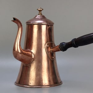 May include: A copper coffee pot with a long, curved spout and a wooden handle. The pot has a lid with a brass finial. The copper has a warm, reflective finish, and the handle is dark brown.