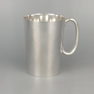 May include: A silver-colored mug with a rounded handle. The mug has a cylindrical body and a simple, elegant design. The interior of the mug reflects light, creating highlights. The mug is likely made of metal.