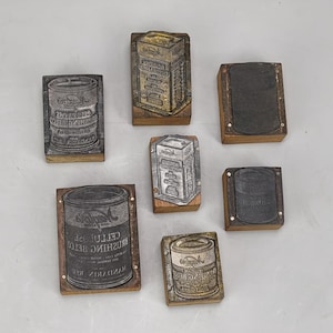 May include: A collection of seven rectangular wooden stamps with metal printing plates. The plates feature engraved illustrations of various cylindrical cans and rectangular boxes, some with visible text including the word Mandarins.