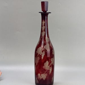 Può includere: Una caraffa alta in vetro rosso scuro con un tappo in vetro trasparente. La caraffa è decorata con un motivo a vite inciso nel vetro.
