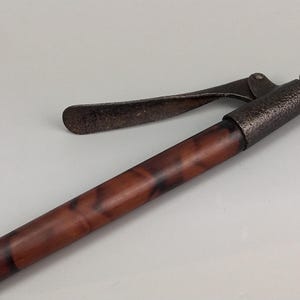 Pode incluir: Uma bengala com um eixo marmoreado em tons de castanho e preto e um punho de metal escuro texturizado. O punho inclui um mecanismo de alavanca curvo e articulado no topo, rematado com uma pequena tampa circular de tom latão na extremidade.