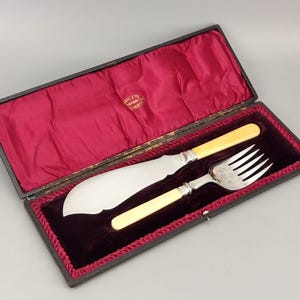 Puede incluir: Un juego de servir pescado vintage en un estuche negro con interior de terciopelo rojo. El juego incluye un cuchillo grande y un tenedor con mangos color marfil y detalles plateados. El estuche está abierto.