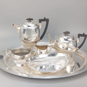 Può includere: Un servizio da tè in argento su un vassoio d'argento. Il set comprende una teiera, una lattiera, una zuccheriera e un piatto da portata. La teiera ha un manico nero e un coperchio decorativo. Il vassoio ha manici su ogni lato.