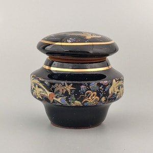 Peut inclure: Un petit pot en céramique noire avec un couvercle, décoré de motifs floraux dorés et colorés.