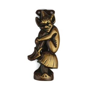 Puede incluir: Un llamador de puerta de bronce con forma de duende sentado en un hongo. El duende tiene una expresión traviesa y se está sujetando la pierna.