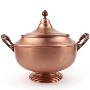 Op de afbeelding: Een koperkleurige serveerschaal met deksel. De kom heeft twee sierlijke handvatten en een decoratieve bekroning op het deksel. De kom staat op een ronde voet en is gemaakt van glanzend metaal.