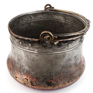 Puede incluir: Un pequeño cubo de metal antiguo con asa. El cubo es plateado con detalles de color cobre y un borde decorativo. El asa y los elementos decorativos son de color dorado. El cubo probablemente se utiliza con fines decorativos.