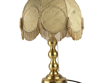 May include: A gold table lamp with a beige fabric lampshade with a beaded fringe. The lampshade has a scalloped design.