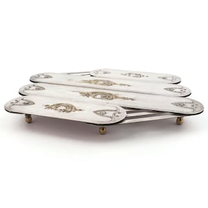 Puede incluir: Salvamanteles plegable plateado con diseños de filigrana dorados ornamentados. El salvamanteles tiene cuatro secciones ovaladas y pequeños pies de color latón. El diseño es simétrico y elegante.