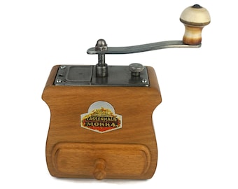 May include: A vintage wooden coffee grinder with a metal crank and a drawer for collecting grounds. The grinder has a label that reads "Lassenhaus Mokka".