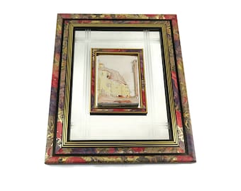 May include: A framed mirror with a smaller framed picture inside. The outer frame is made of wood with a floral pattern in shades of red, gold, and black. The inner picture is a watercolour painting of a building with a yellow facade.